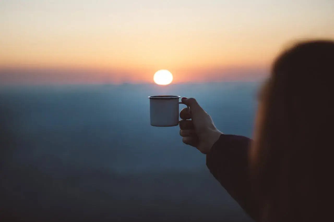 Une tasse de cafée brandie à bout de bras au-dessus des montagnes, en face d'un soleil couchant.